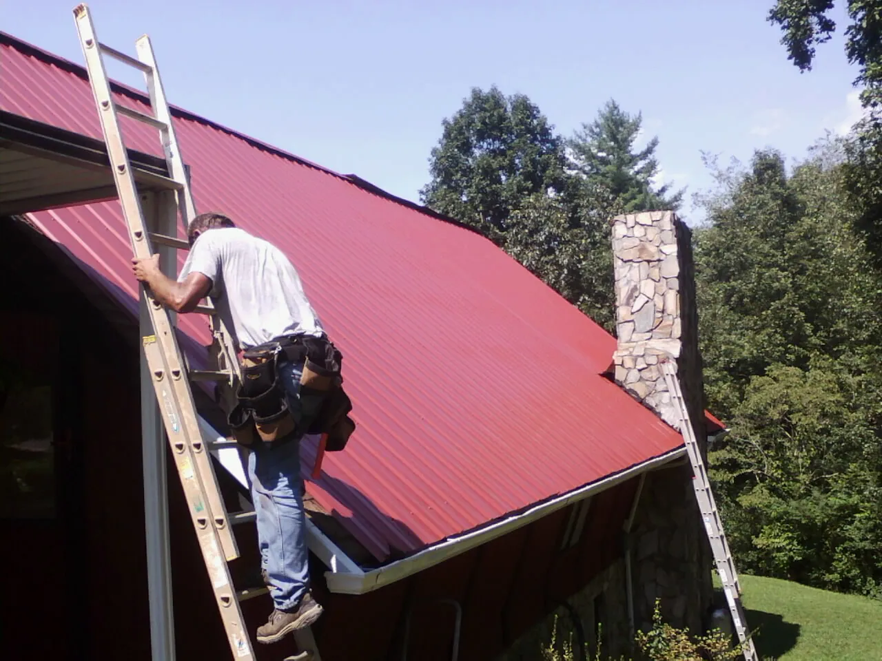 Roofer on a ladder beside a red metal roof and stone chimney — Soffit & Fascia Repair by Roofing Brothers in Medinah